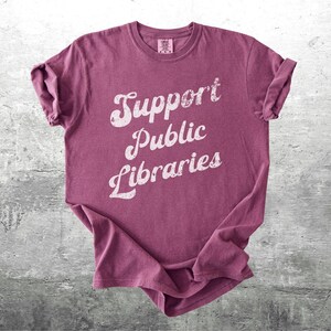 May include: A maroon t-shirt with the words "Support Public Libraries" in a distressed white font. The shirt is folded and laid on a gray, textured surface. The sleeves are cuffed.