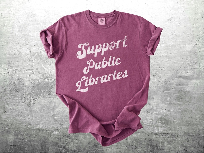 May include: A maroon t-shirt with the words "Support Public Libraries" in a distressed white font. The shirt is folded and laid on a gray, textured surface. The sleeves are cuffed.