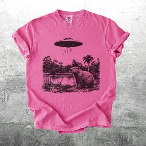 May include: A pink t-shirt featuring a black and white graphic of a capybara standing near a body of water with a UFO hovering above. The design includes palm trees and other foliage. The shirt has rolled-up sleeves.