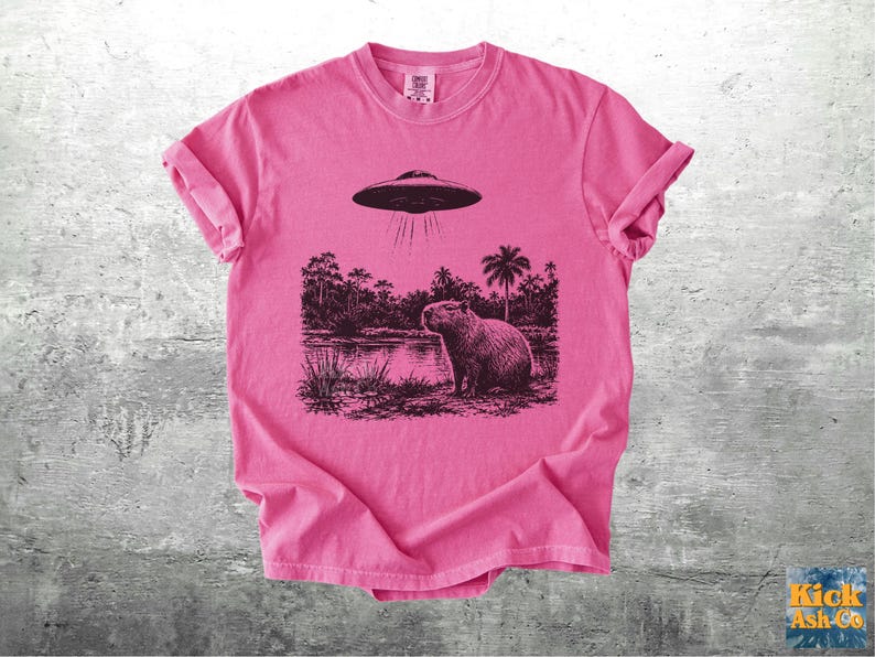 May include: A pink t-shirt featuring a black and white graphic of a capybara standing near a body of water with a UFO hovering above. The design includes palm trees and other foliage. The shirt has rolled-up sleeves.