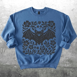 May include: A blue sweatshirt featuring a black bat design with outstretched wings, surrounded by floral and leaf patterns. The bat has a detailed face and is centered on the front of the garment. The sleeves are rolled up.