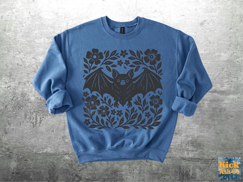 May include: A blue sweatshirt featuring a black bat design with outstretched wings, surrounded by floral and leaf patterns. The bat has a detailed face and is centered on the front of the garment. The sleeves are rolled up.