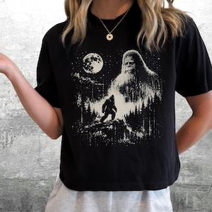 以下が含まれることがあります： 黒の半袖Tシャツ。満月と星空の下、森の中で小さなビッグフットのシルエットを見下ろす大きなビッグフットの姿が、かすれた質感の白いグラフィックプリントで描かれています。