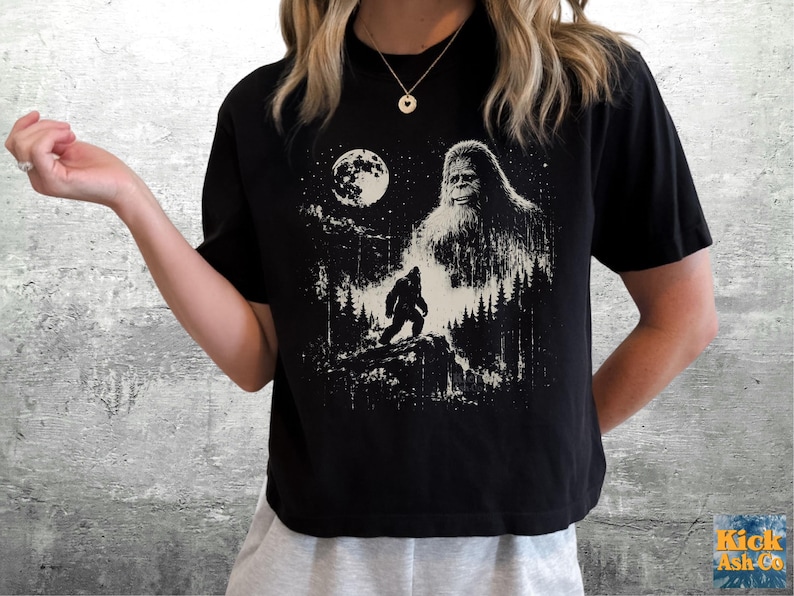 以下が含まれることがあります： 黒の半袖Tシャツ。満月と星空の下、森の中で小さなビッグフットのシルエットを見下ろす大きなビッグフットの姿が、かすれた質感の白いグラフィックプリントで描かれています。
