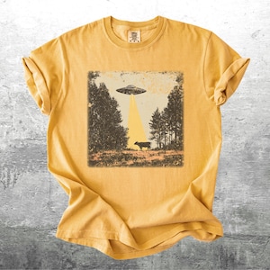 May include: Mustard yellow t-shirt featuring a distressed graphic of a UFO beaming a cow. The design includes trees and a beam of light. The shirt is made of soft material and has a classic crew neck.