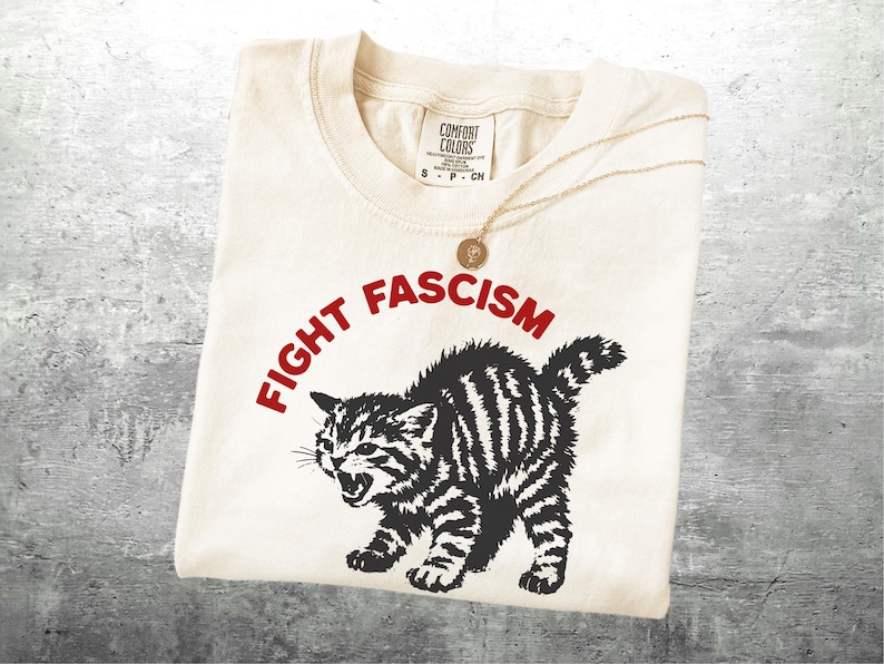 K&ouml;nnte beinhalten: Cremefarbenes T-Shirt mit dem roten Schriftzug "FIGHT FASCISM" &uuml;ber einer schwarz-wei&szlig;en Illustration einer w&uuml;tenden Katze. Eine goldene Halskette mit Anh&auml;nger liegt auf dem Shirt.
