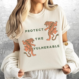 May include: A cream colored t-shirt with a graphic of two orange tigers. The text "PROTECT THE VULNERABLE" is printed in green on the shirt.