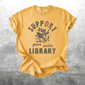 May include: Mustard yellow t-shirt with rolled sleeves. The front features the text "SUPPORT your public LIBRARY" in a distressed font, with a bird perched on a flowering branch graphic above the words.