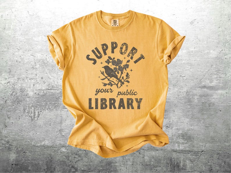May include: Mustard yellow t-shirt with rolled sleeves. The front features the text "SUPPORT your public LIBRARY" in a distressed font, with a bird perched on a flowering branch graphic above the words.