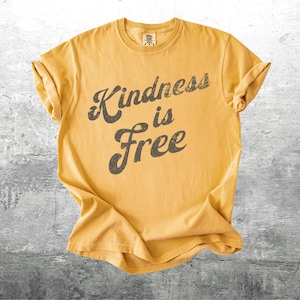 May include: Mustard yellow t-shirt with rolled sleeves. The shirt features the phrase "Kindness is Free" in a distressed, dark gray font. The shirt is laid flat on a gray, textured surface.