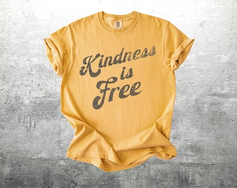 May include: Mustard yellow t-shirt with rolled sleeves. The shirt features the phrase "Kindness is Free" in a distressed, dark gray font. The shirt is laid flat on a gray, textured surface.