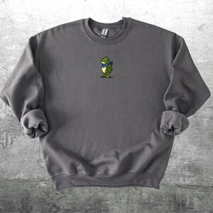 Puede incluir: Sudadera gris carb&oacute;n con un peque&ntilde;o bordado de una rana con una bufanda azul. La rana es verde con detalles amarillos. La sudadera tiene mangas largas y cuello redondo. La tela parece suave y c&oacute;moda.