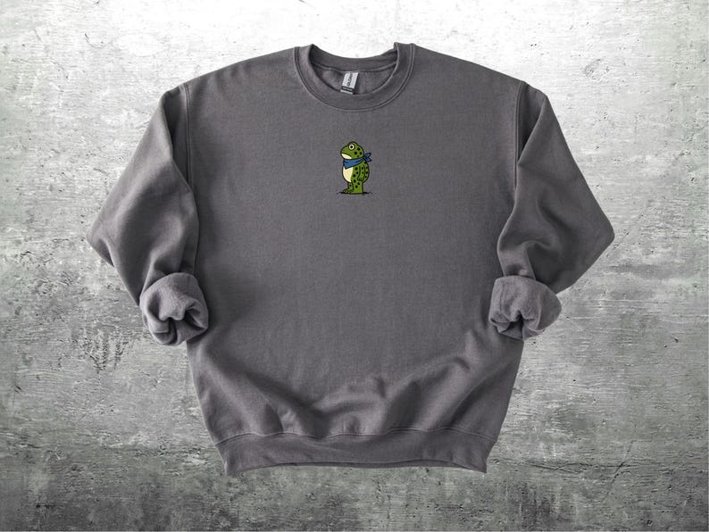 Puede incluir: Sudadera gris carb&oacute;n con un peque&ntilde;o bordado de una rana con una bufanda azul. La rana es verde con detalles amarillos. La sudadera tiene mangas largas y cuello redondo. La tela parece suave y c&oacute;moda.