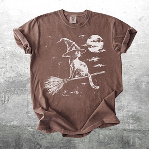 May include: A brown short-sleeve t-shirt featuring a white distressed graphic print of a cat wearing a pointed witch hat, sitting on a flying broomstick in front of a full moon and clouds. The shirt has a crew neckline and a relaxed fit.