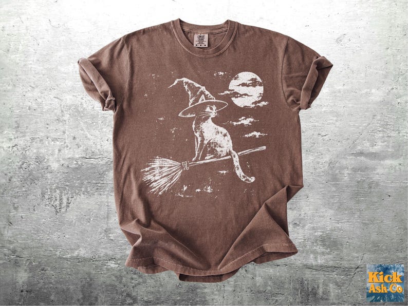 May include: A brown short-sleeve t-shirt featuring a white distressed graphic print of a cat wearing a pointed witch hat, sitting on a flying broomstick in front of a full moon and clouds. The shirt has a crew neckline and a relaxed fit.
