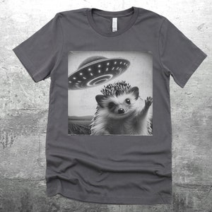 May include: A dark gray t-shirt with a black and white image of a hedgehog waving at a flying saucer.