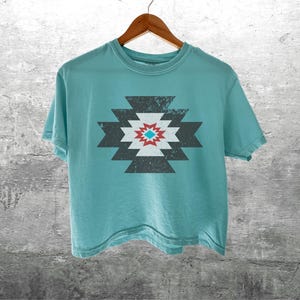 May include: A turquoise t-shirt with a geometric design in the center. The design features a series of dark gray, white, and red shapes arranged in a symmetrical pattern. The shirt is hanging on a wooden hanger against a gray concrete wall.