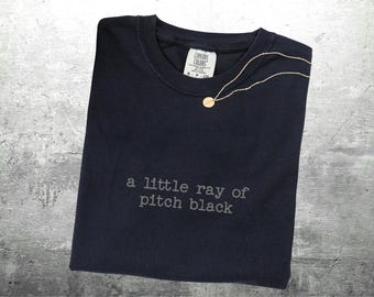 May include: A navy blue t-shirt with the phrase "a little ray of pitch black" printed in gray. A gold-colored necklace with a small circular pendant rests on the shirt. The shirt is folded and lies on a gray surface.