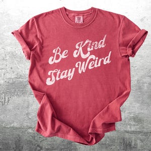 May include: A red t-shirt with the words "Be Kind Stay Weird" in a white, cursive font. The shirt is folded and has rolled-up sleeves. The background is a textured, gray surface.