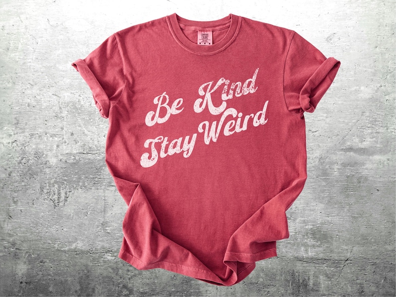 May include: A red t-shirt with the words "Be Kind Stay Weird" in a white, cursive font. The shirt is folded and has rolled-up sleeves. The background is a textured, gray surface.