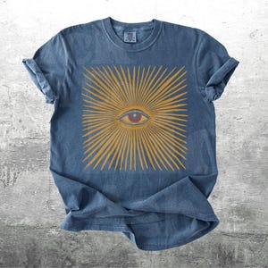 May include: A blue t-shirt featuring a gold sunburst design with a central eye. The eye is red and the rays of the sunburst are gold. The shirt has rolled-up sleeves and a crew neck.