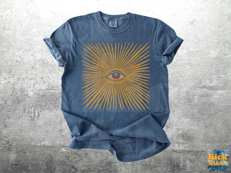 May include: A blue t-shirt featuring a gold sunburst design with a central eye. The eye is red and the rays of the sunburst are gold. The shirt has rolled-up sleeves and a crew neck.