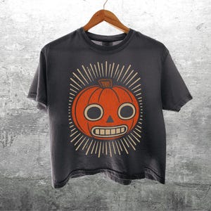 May include: A charcoal gray short-sleeve t-shirt featuring a central graphic of an orange pumpkin with a stylized face, round eyes, a triangular nose, and a rectangular toothy grin, surrounded by radiating light lines on a dark background.