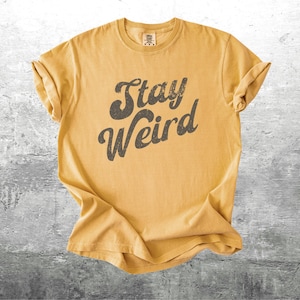 K&ouml;nnte beinhalten: Senfgelbes T-Shirt mit hochgekrempelten &Auml;rmeln. Das Shirt zeigt den Schriftzug "Stay Weird" in einer dunkelgrauen Schrift im Retro-Stil. Das Shirt ist aus einem weichen, bequemen Material gefertigt.
