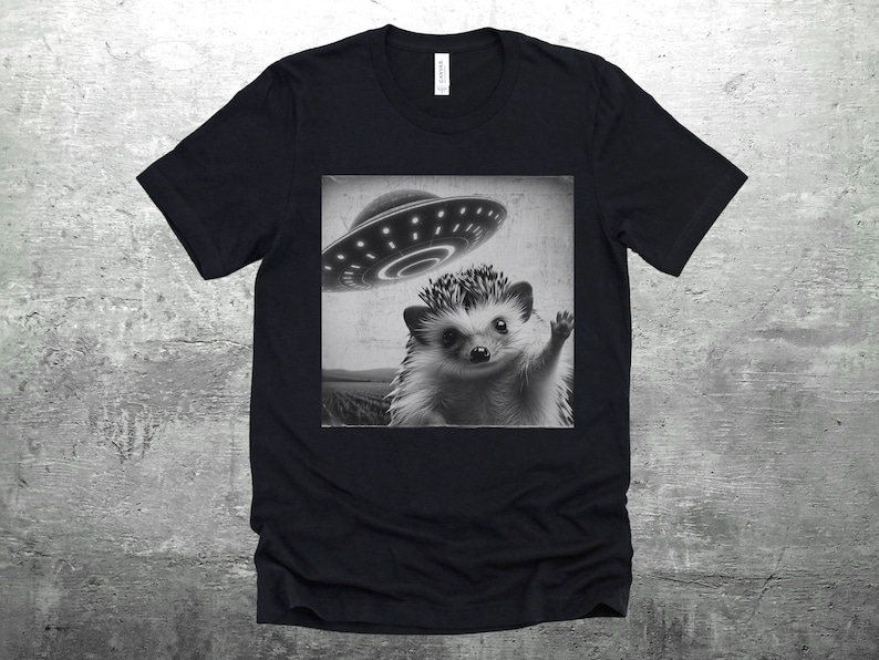 May include: Black t-shirt with a graphic of a hedgehog wearing a UFO hat. The hedgehog is looking at the camera and waving.