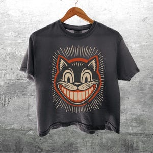 May include: A charcoal gray short-sleeve t-shirt featuring a graphic print of a grinning cat face with large eyes and a wide smile, surrounded by a sunburst pattern in orange and cream colors. The shirt hangs on a wooden hanger.