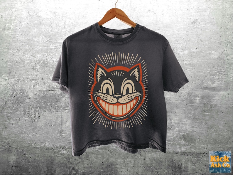 May include: A charcoal gray short-sleeve t-shirt featuring a graphic print of a grinning cat face with large eyes and a wide smile, surrounded by a sunburst pattern in orange and cream colors. The shirt hangs on a wooden hanger.