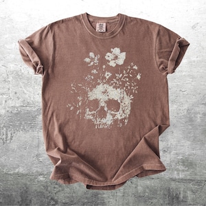 Pu&ograve; includere: T-shirt marrone con un design a teschio invecchiato con dettagli floreali. La grafica &egrave; in bianco sbiadito, creando un'estetica vintage. La maglietta ha un girocollo e maniche corte.