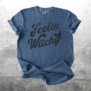 K&ouml;nnte beinhalten: Ein blaues T-Shirt mit hochgekrempelten &Auml;rmeln. Das Shirt zeigt den Schriftzug "Feelin Witchy" in Schwarz, mit einer Hexenhut-Grafik. Das T-Shirt ist aus weichem Material und hat einen Vintage-Look.