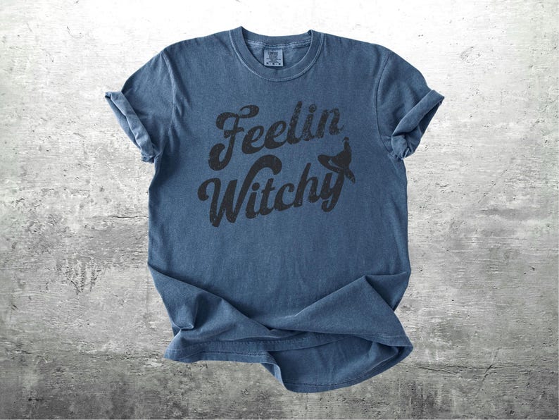 K&ouml;nnte beinhalten: Ein blaues T-Shirt mit hochgekrempelten &Auml;rmeln. Das Shirt zeigt den Schriftzug "Feelin Witchy" in Schwarz, mit einer Hexenhut-Grafik. Das T-Shirt ist aus weichem Material und hat einen Vintage-Look.