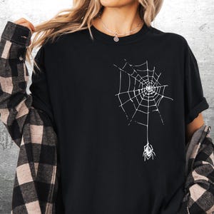 May include: Black t-shirt featuring a white spider web design with a spider hanging below. The shirt is a solid black color. The person is wearing a plaid shirt over the black t-shirt.
