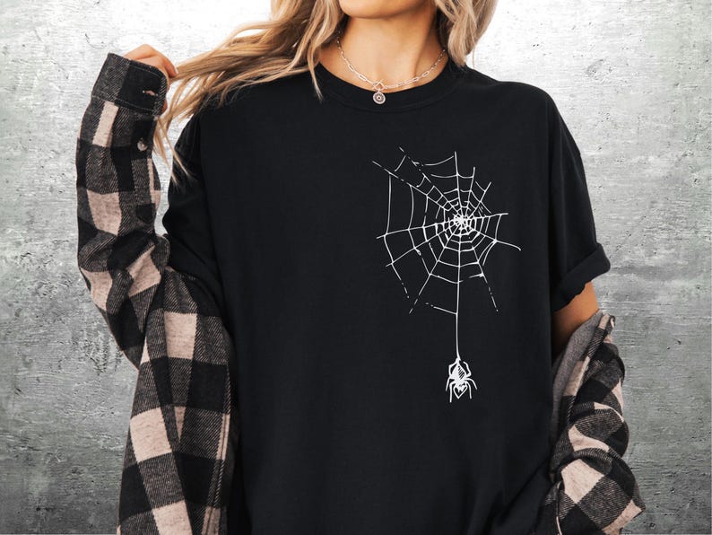 May include: Black t-shirt featuring a white spider web design with a spider hanging below. The shirt is a solid black color. The person is wearing a plaid shirt over the black t-shirt.