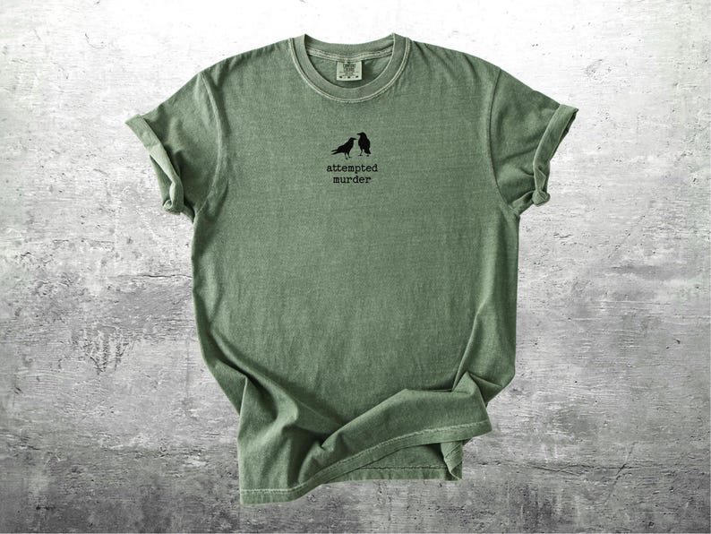 May include: Sage green t-shirt with rolled sleeves. The shirt features a small black graphic of two birds above the words "attempted murder" in a simple font. The shirt is made of a soft material.