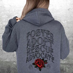 Pu&ograve; includere: Una felpa con cappuccio grigio scuro con il testo "FOSTER ADOPT RESCUE ADVOCATE" stampato in nero sul retro. Una rosa rossa con foglie verdi e due impronte di zampa si trovano sotto il testo.