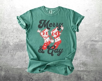May include: Teal t-shirt with the text "Merry & Gay" in a vintage style. The graphic shows two cartoon Christmas stockings with smiling faces and red polka dots. A cheerful and festive design.
