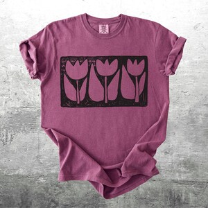 May include: A maroon t-shirt featuring a black graphic of three stylized tulips. The design is rectangular and centered on the shirt. The shirt has rolled-up sleeves and is displayed on a gray, textured surface.
