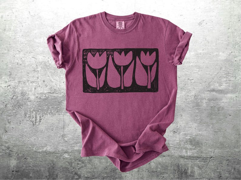 May include: A maroon t-shirt featuring a black graphic of three stylized tulips. The design is rectangular and centered on the shirt. The shirt has rolled-up sleeves and is displayed on a gray, textured surface.