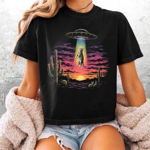 May include: A black t-shirt featuring a graphic print of a tabby cat being abducted by a UFO in a desert landscape. The scene includes cacti, a sunset with purple and orange clouds, and a bright blue tractor beam lifting the cat into the air.