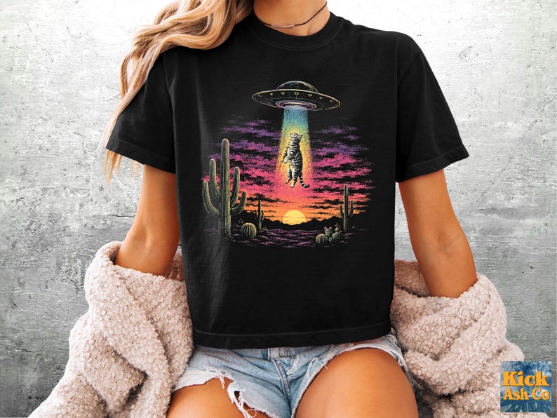 May include: A black t-shirt featuring a graphic print of a tabby cat being abducted by a UFO in a desert landscape. The scene includes cacti, a sunset with purple and orange clouds, and a bright blue tractor beam lifting the cat into the air.