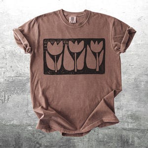May include: A brown t-shirt with a black graphic of three stylized tulips in a rectangular frame. The shirt has a crew neck and short sleeves, with the sleeves rolled up. The fabric has a slightly faded appearance.