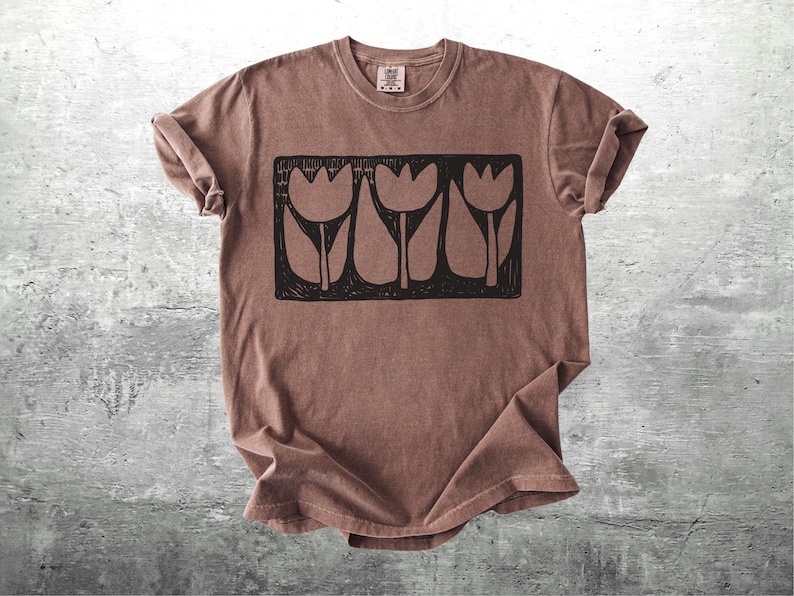 May include: A brown t-shirt with a black graphic of three stylized tulips in a rectangular frame. The shirt has a crew neck and short sleeves, with the sleeves rolled up. The fabric has a slightly faded appearance.