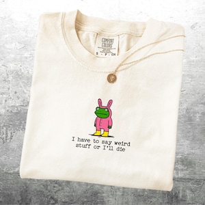May include: Cream-coloured t-shirt featuring a cartoon frog in a pink bunny suit and yellow boots. The text "I have to say weird stuff or I'll die" is printed below the frog. A gold necklace with a pendant is draped over the shirt.
