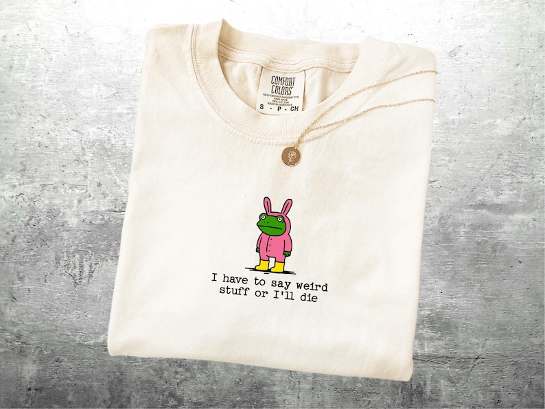 May include: Cream-coloured t-shirt featuring a cartoon frog in a pink bunny suit and yellow boots. The text "I have to say weird stuff or I'll die" is printed below the frog. A gold necklace with a pendant is draped over the shirt.