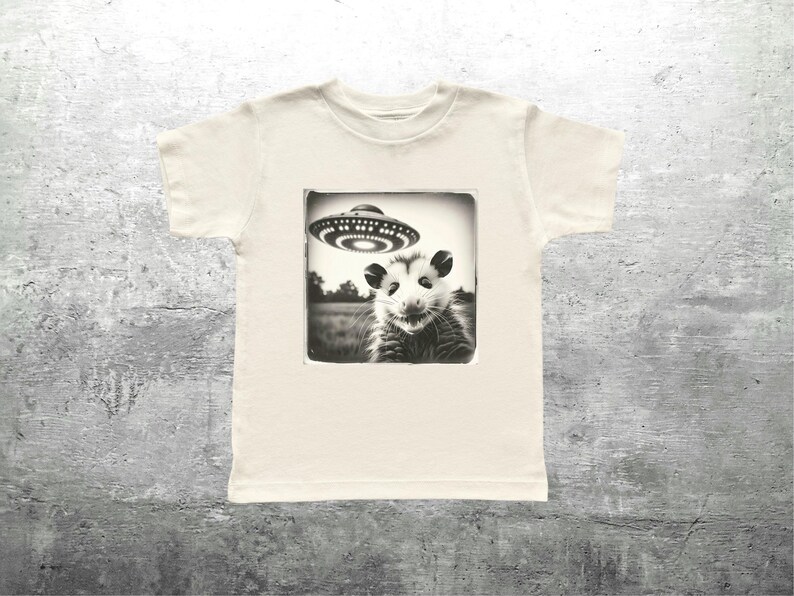 May include: Off-white t-shirt with a black and white image of an opossum and a UFO. The opossum is in a field, and the UFO is above it. The shirt is made of a soft material.