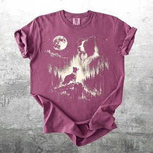 Pu&ograve; includere: T-shirt bordeaux con grafica di un cane sotto la luna piena e le stelle. Il design presenta una sagoma di cane, la testa di un cane pi&ugrave; grande e un cane pi&ugrave; piccolo su una collina, con alberi sullo sfondo.