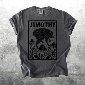 Peut inclure: Un t-shirt gris anthracite &agrave; manches courtes avec une impression graphique noire repr&eacute;sentant un raton laveur sautant au-dessus de grandes fleurs. Le texte JIMOTHY est imprim&eacute; en gras, en noir, au sommet du cadre rectangulaire central.